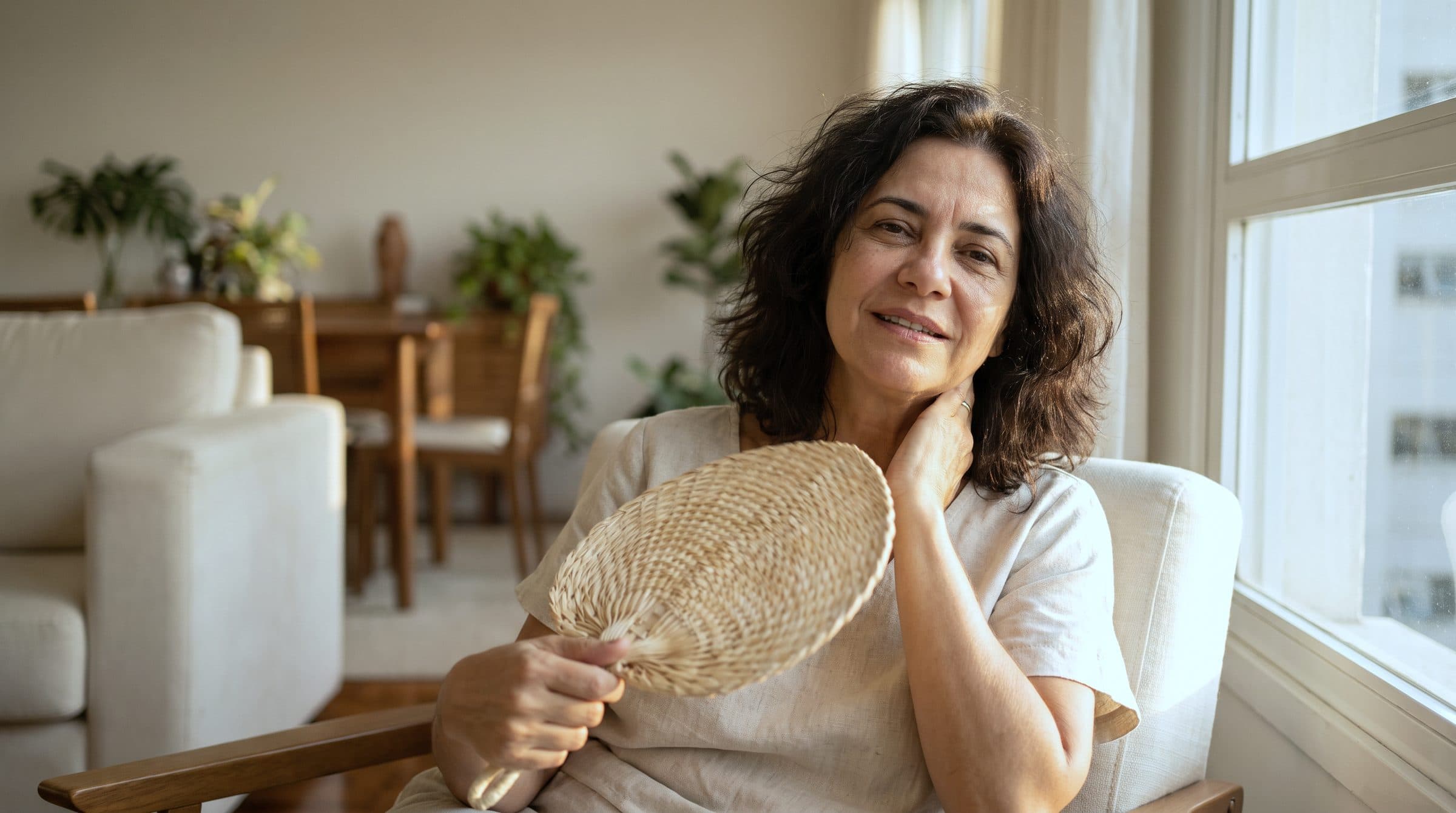 Mulher de cerca de 50 anos se abanando com um leque de palha na sala, no fim da tarde
